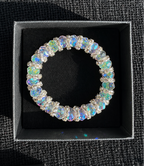 A silver-toned bracelet adorned with iridescent, rainbow-flecked oval crystals, presented inside a black jewelry box on a dark textured fabric background.