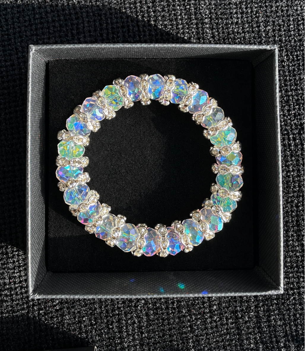 A silver-toned bracelet adorned with iridescent, rainbow-flecked oval crystals, presented inside a black jewelry box on a dark textured fabric background.