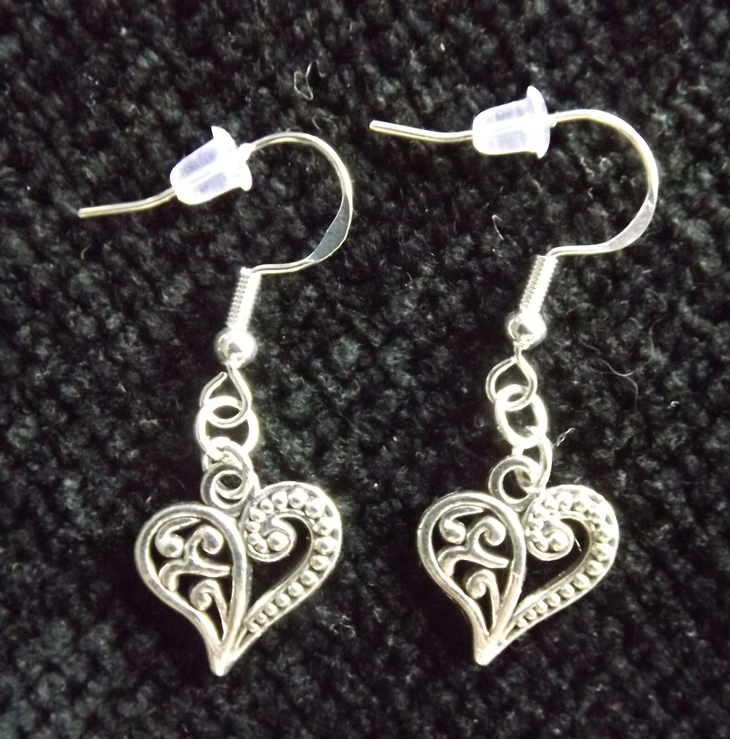 A pair of silver filigree heart dangle earrings with fishhook backings, displayed on a textured black fabric background.