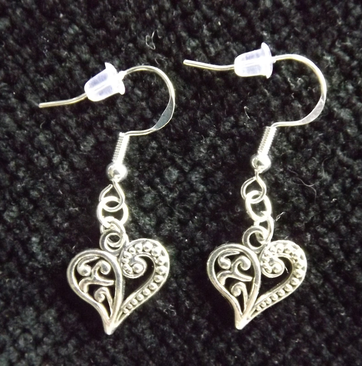 A pair of silver filigree heart dangle earrings with fishhook backings, displayed on a textured black fabric background.