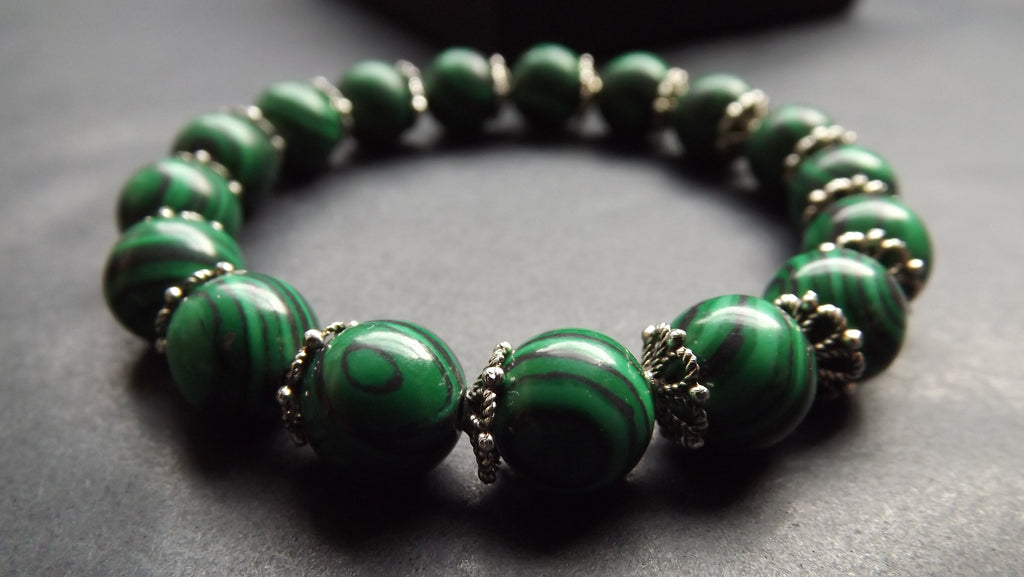 A beaded stretch bracelet made of polished green malachite beads with silver-toned spacer details, shown on a dark gray textured surface.