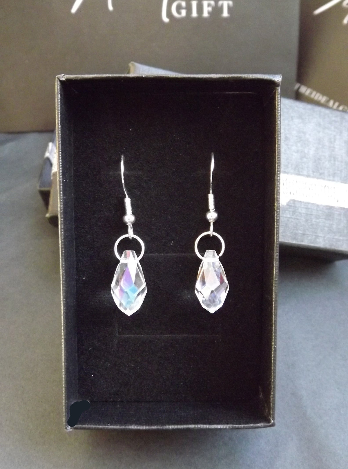 A pair of silver-tone dangle earrings with faceted clear teardrop-shaped crystals, presented in a black gift box. The background shows a dark gray surface and a branded box for 'The Ideal Gift'.