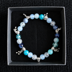 A beaded stretch bracelet with light blue, turquoise, and white beads, silver-toned charms, and clear faceted beads, displayed inside a black presentation box on a textured black background.