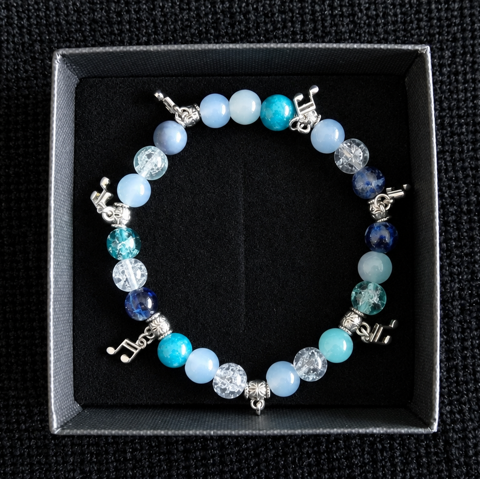 A beaded stretch bracelet with light blue, turquoise, and white beads, silver-toned charms, and clear faceted beads, displayed inside a black presentation box on a textured black background.