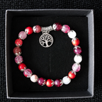 A beaded bracelet with red, pink, and white marbled beads and a silver tree of life charm, displayed inside a black presentation box on a black textured background.