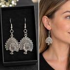 Silver-toned filigree peacock drop earrings displayed in a black presentation box, with a side-by-side close-up of the earrings worn on a woman's ear.