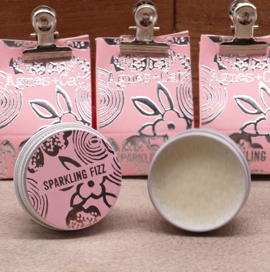 Two tins of lip balm, with pink lids labeled 'Sparkling Fizz' and 'Agnes + Cat' branding, with pink boxes in the background.